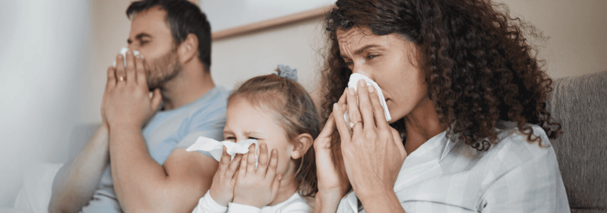 A family sitting on a couch, all using tissues to blow their noses, appearing unwell.